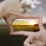 Hands framing sunset over field with hay bales